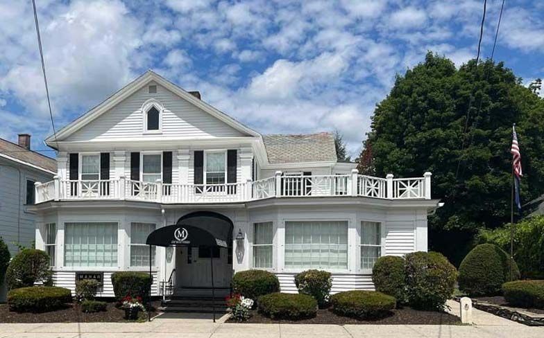 White building with black shutters and a black awning. Bushes and a cloudy sky visible.