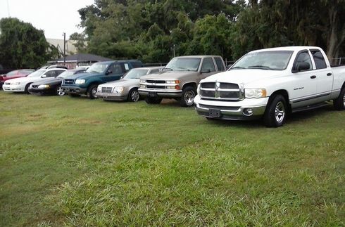 Parked cars and pickup trucks lined up on a grassy lot, with a white pickup in the foreground.
