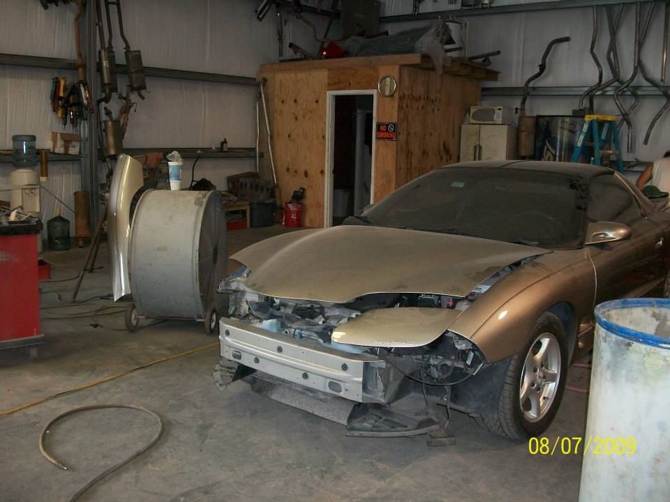 Dusty tan sports car in a garage with its front bumper removed and a wooden enclosure behind it.