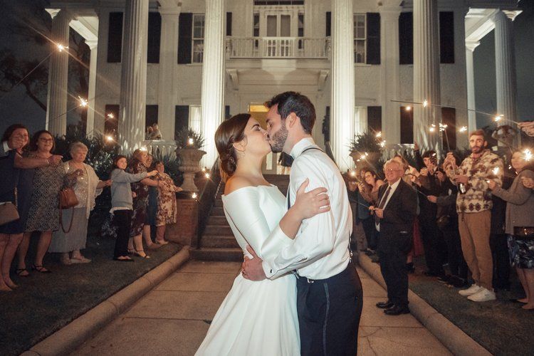 A bride and groom are kissing in front of a large building while their guests hold sparklers.