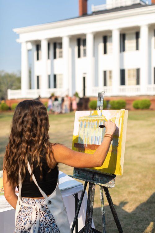 A woman is painting on an easel in front of a large white building.