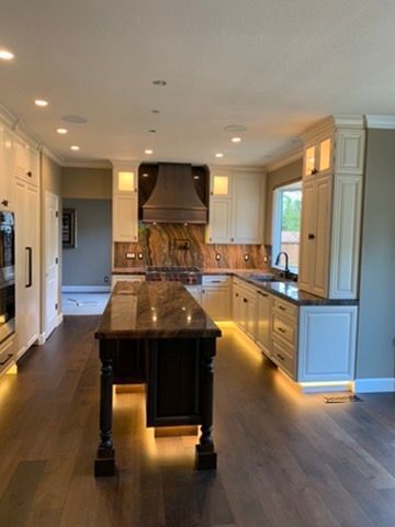 A modern kitchen featuring a dark wood island, light cabinets, stone countertops, a large hood vent, and under-cabinet glow.