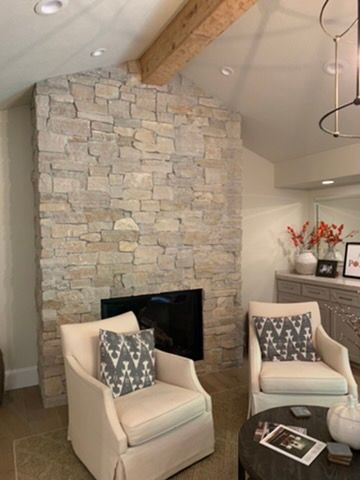 A fireplace with a stone facade, a wooden ceiling beam, and two cream-colored armchairs with patterned pillows.