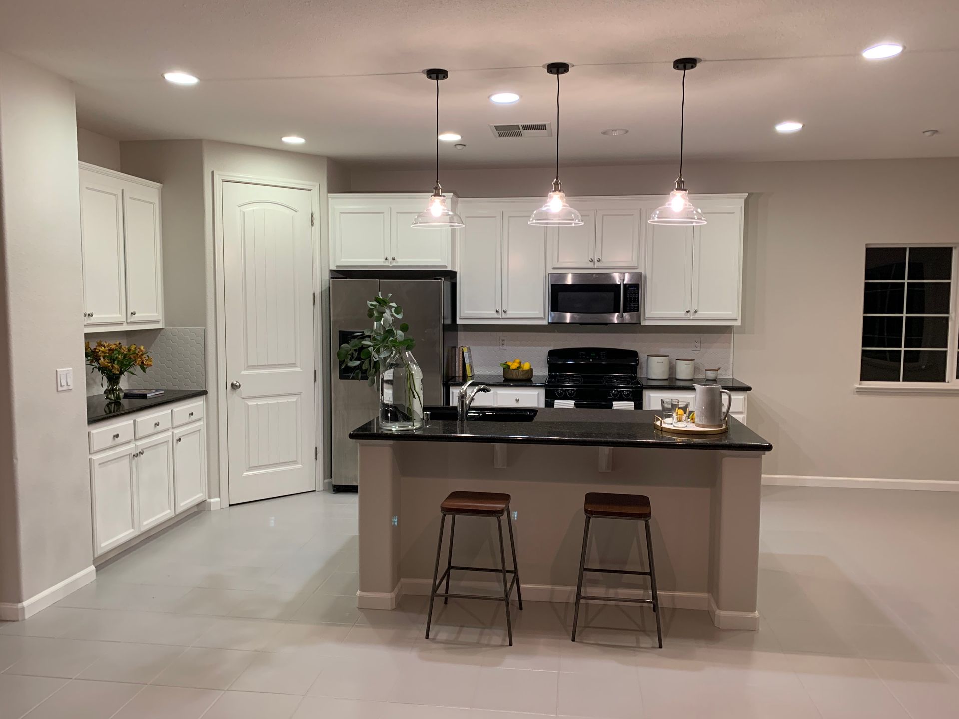 A bright, modern kitchen features white cabinetry, black countertops, a central island with two stools, and hanging lights.