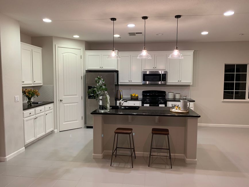 A bright, modern kitchen features white cabinetry, black countertops, a central island with two stools, and hanging lights.