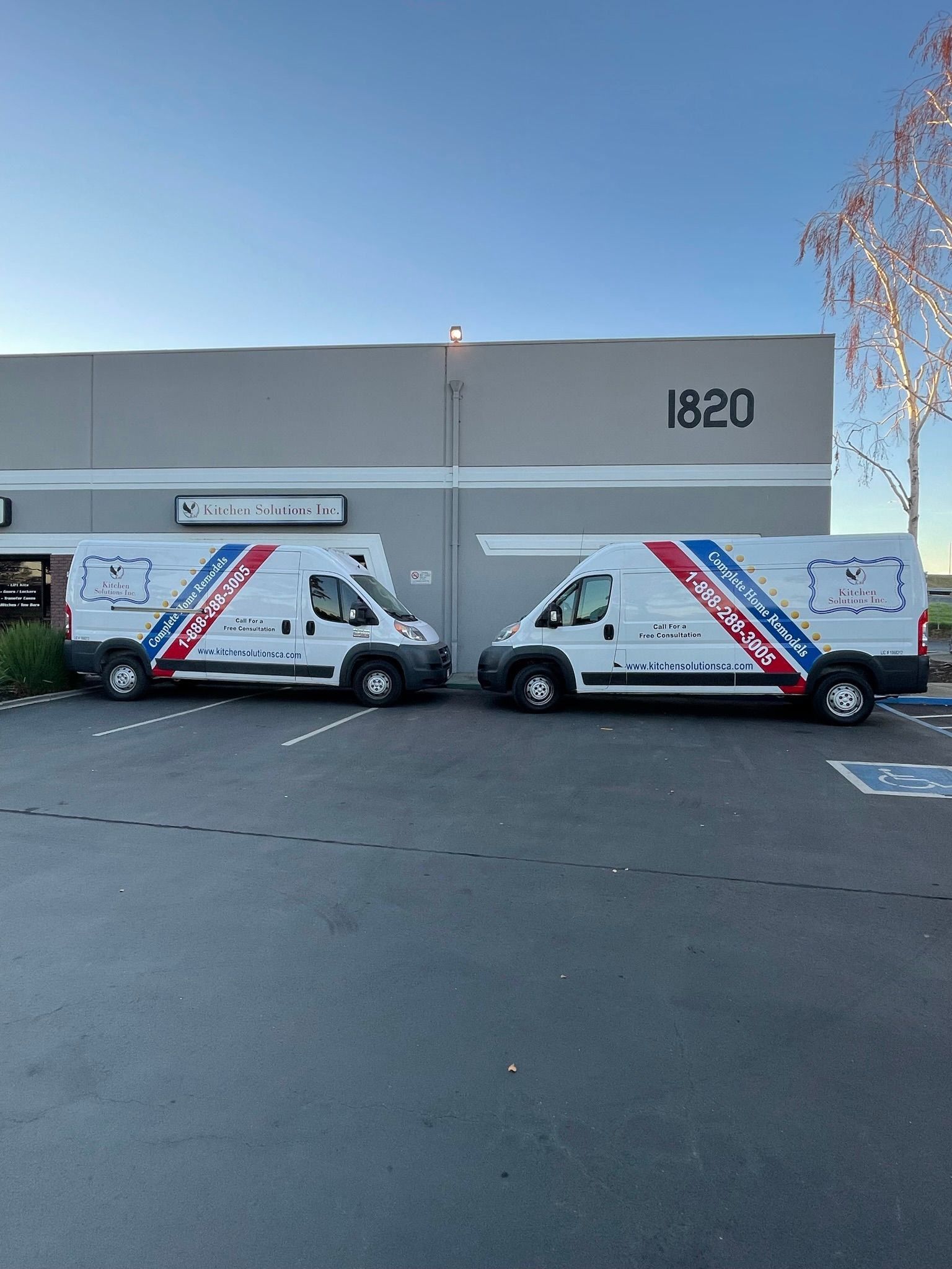 Two white service vans with blue and red side decals parked in front of a building numbered 1820 under a clear blue sky.