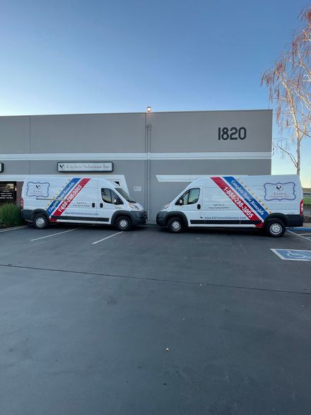 Two white service vans with blue and red side decals parked in front of a building numbered 1820 under a clear blue sky.
