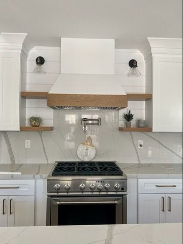 Modern white kitchen with a stainless steel range, marble backsplash, floating wood shelves, and a white vent hood.