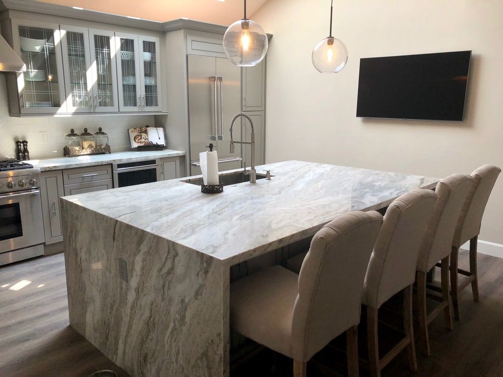 Modern kitchen with a large marble-topped island, four upholstered chairs, stainless appliances, and hanging glass lights.