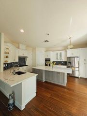 A kitchen with white cabinets, a grey island, marble countertops, stainless steel appliances, and wood floors.