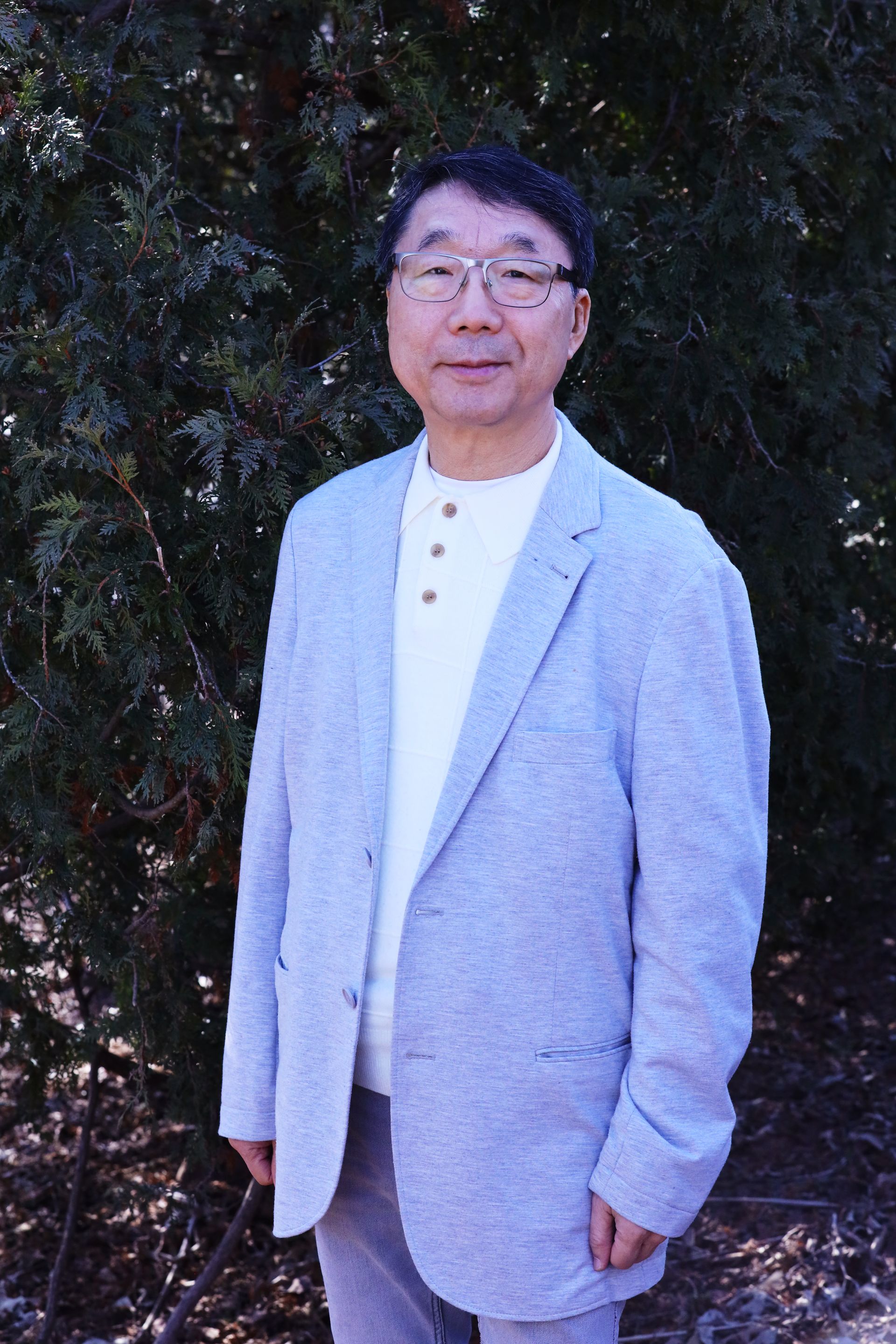 A man in a suit and glasses is standing in front of a tree.