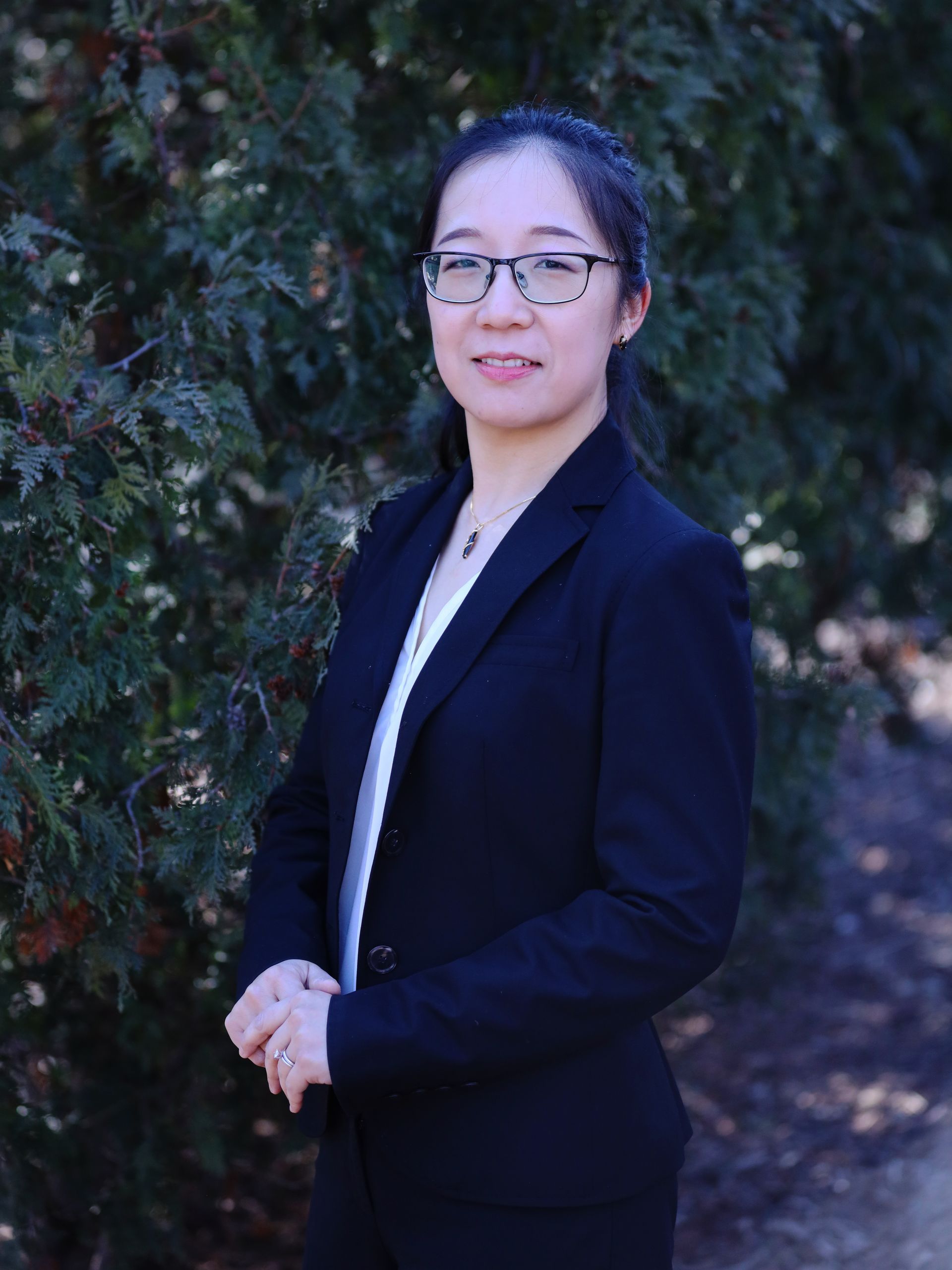 A woman in a suit and glasses is standing in front of a tree.