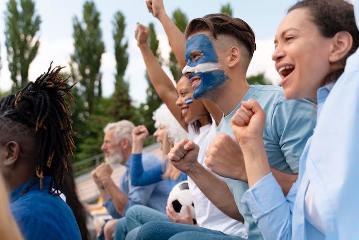 A group of excited sports fans cheering with arms raised at an outdoor stadium, one fan has face paint on.