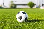 A black and white soccer ball sits on a vibrant green grass field with a blurred building in the background.