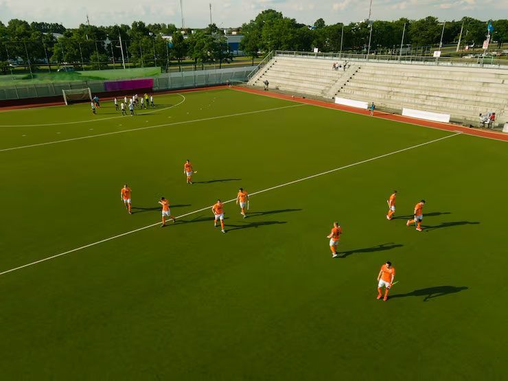 Field hockey players in orange uniforms practice on a green field in a stadium with large concrete bleachers.