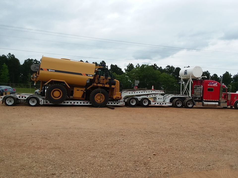 A red semi truck is carrying a large yellow tractor on a trailer.