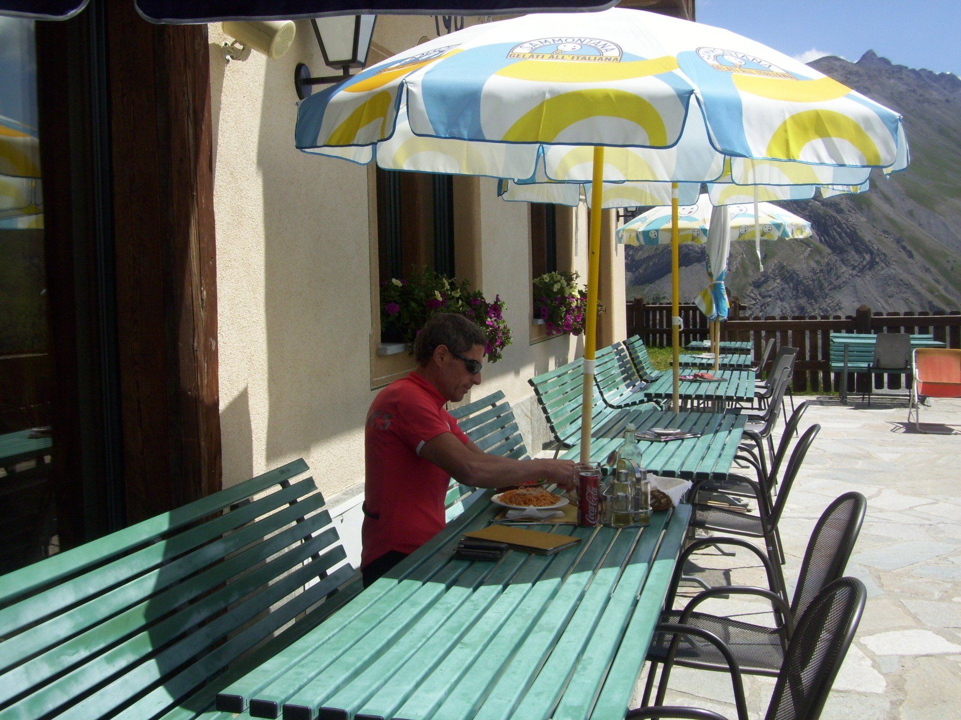 Persona seduta a un tavolo con del cibo sotto un ombrellone nel patio di un ristorante con le montagne sullo sfondo.