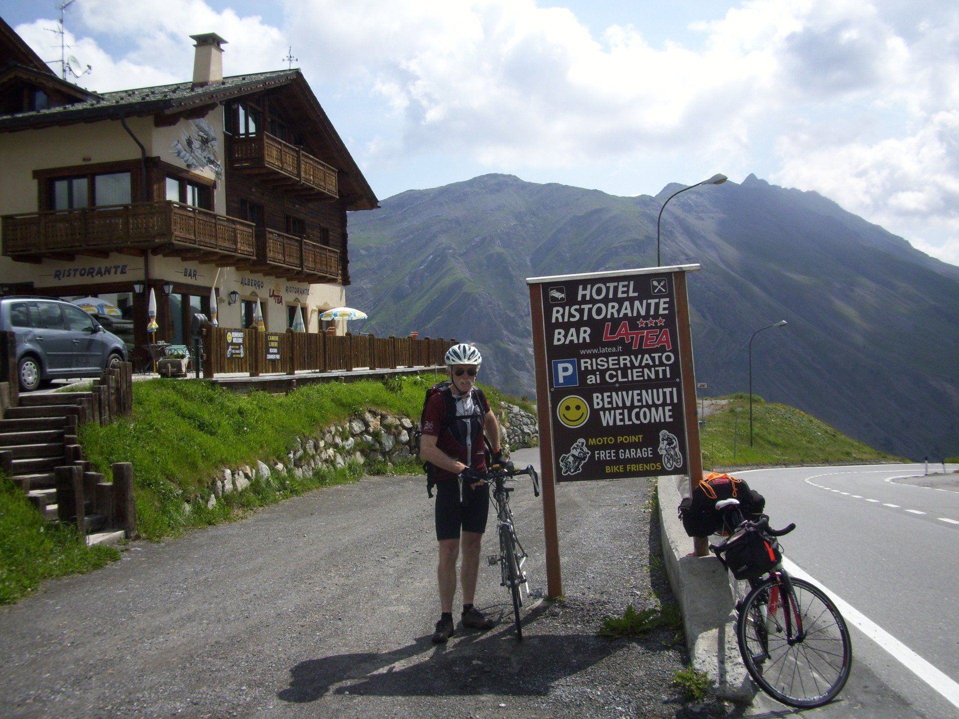Ciclista con la bicicletta in piedi accanto a un cartello di fronte a un hotel di montagna. Montagne sullo sfondo.