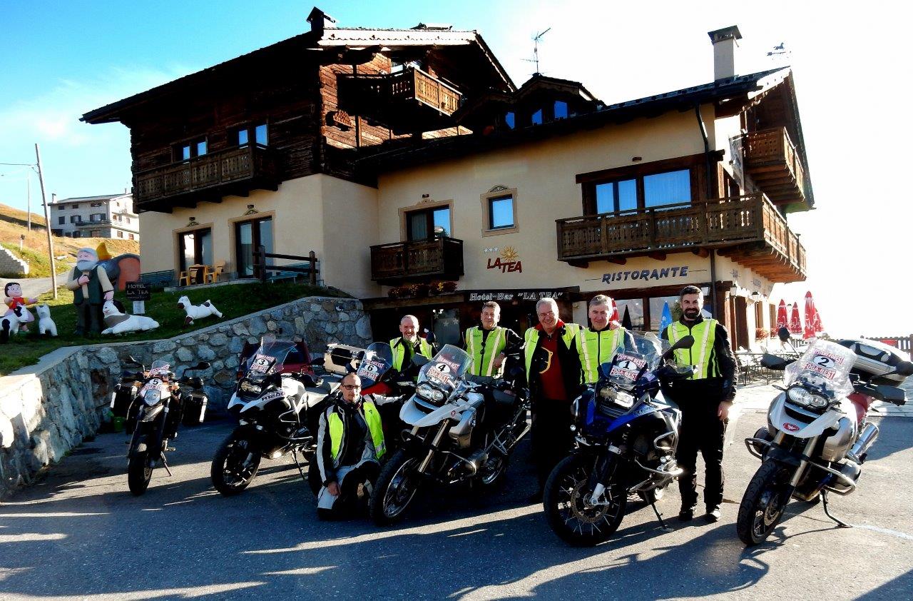 Gruppo di motociclisti in posa davanti a un ristorante in montagna, giornata di sole.