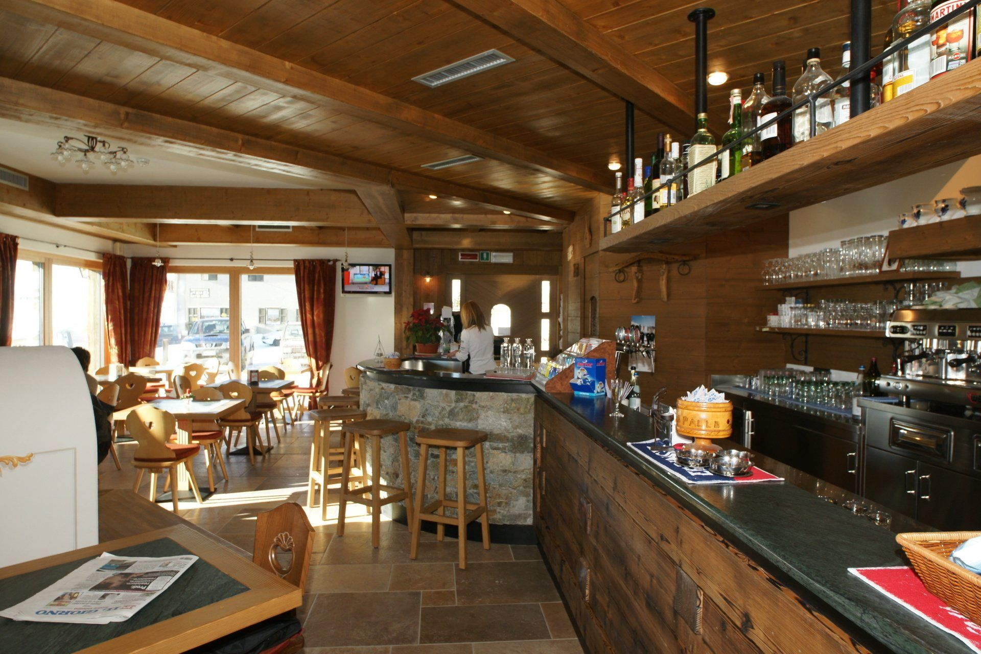 Interno di un bar in legno con un lungo bancone, sgabelli e una piccola zona pranzo con tavoli e sedie.