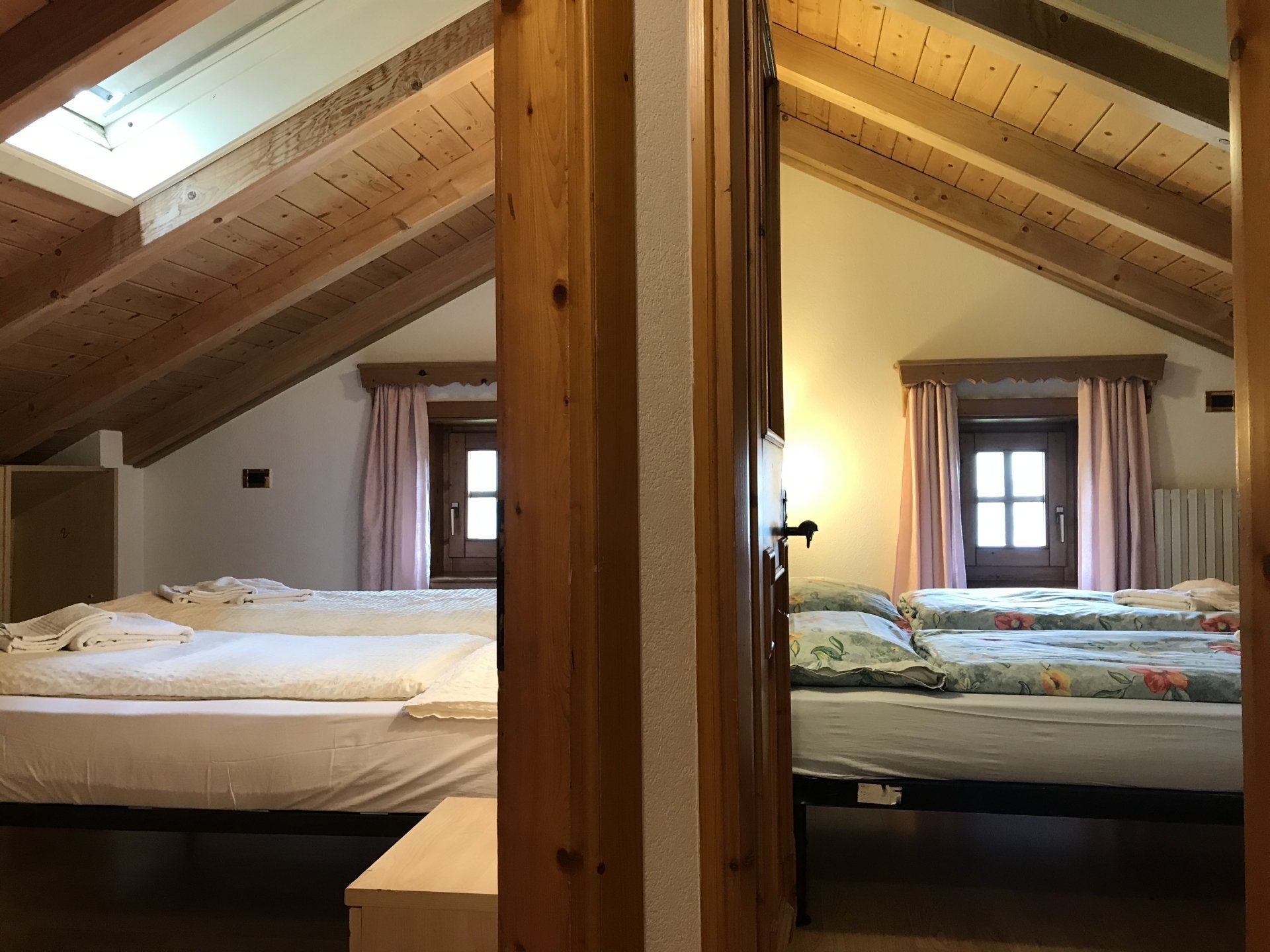 Due camere da letto con letti, visibili attraverso una porta aperta; soffitti in legno, finestre e tende rosa.
