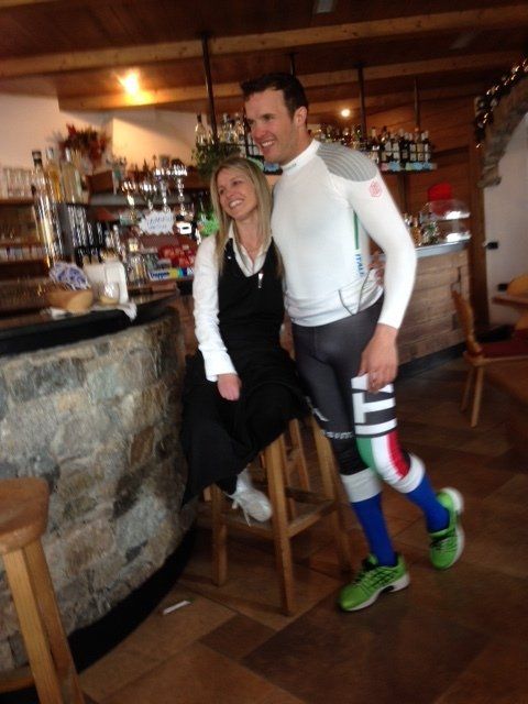 Donna e uomo in posa vicino a un bar. L'uomo è vestito di bianco con indumenti a compressione, la donna con un grembiule nero. Ambiente da bar.