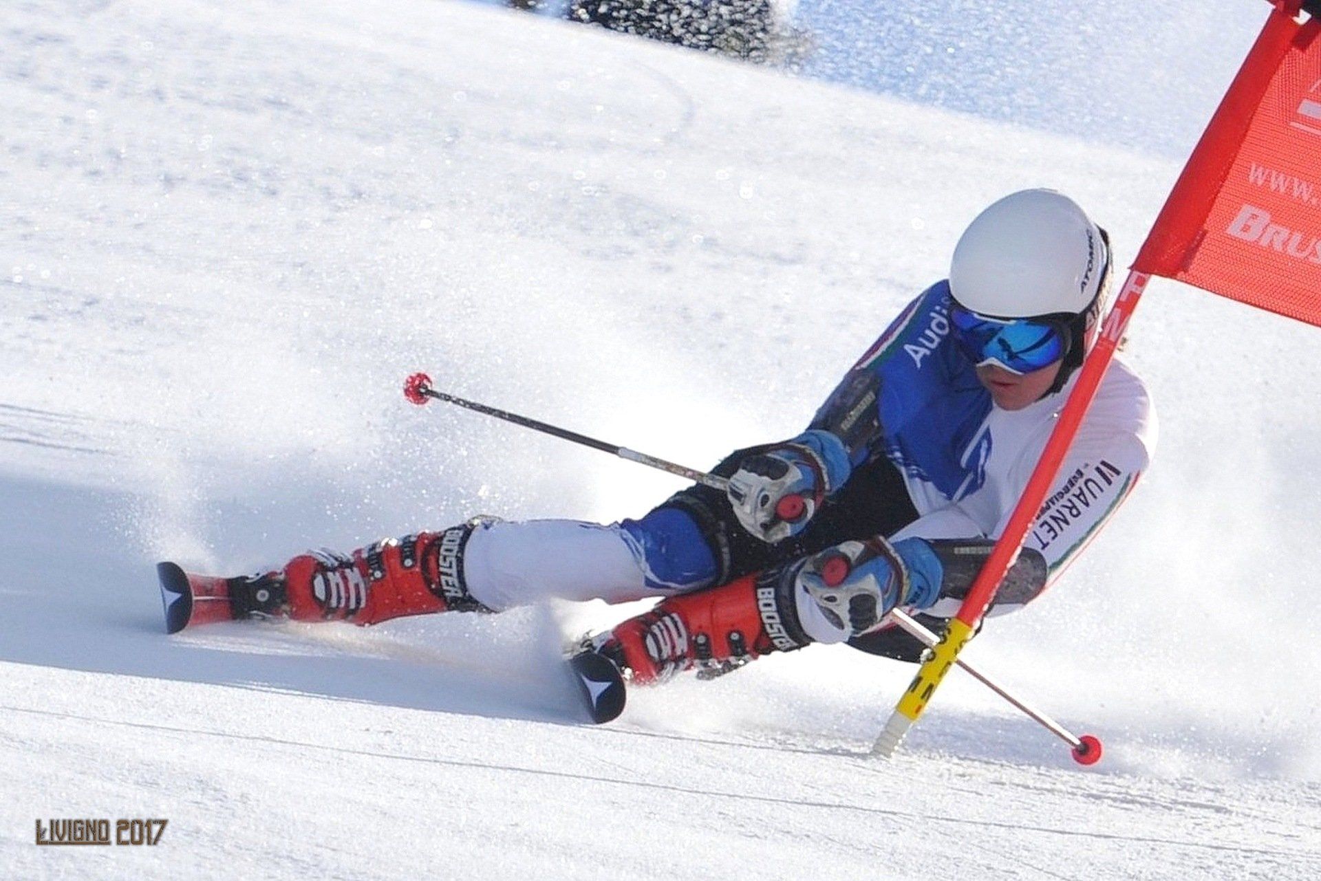Uno sciatore con casco bianco e tuta blu e bianca compie una curva brusca attorno a un cancello rosso su un pendio innevato.