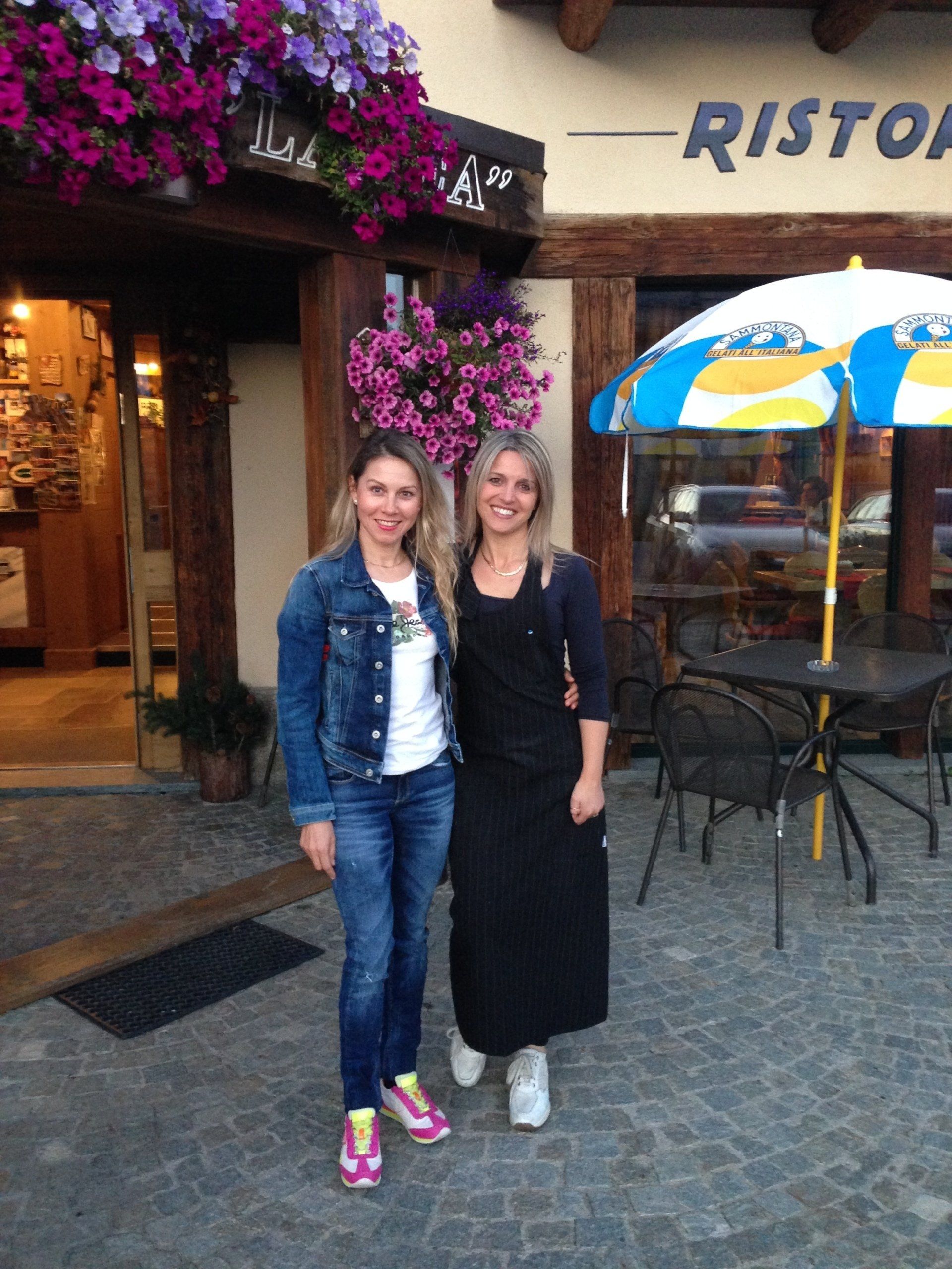 Due donne sorridono fuori da un ristorante con delle fioriere, una in jeans, l'altra con un grembiule nero.