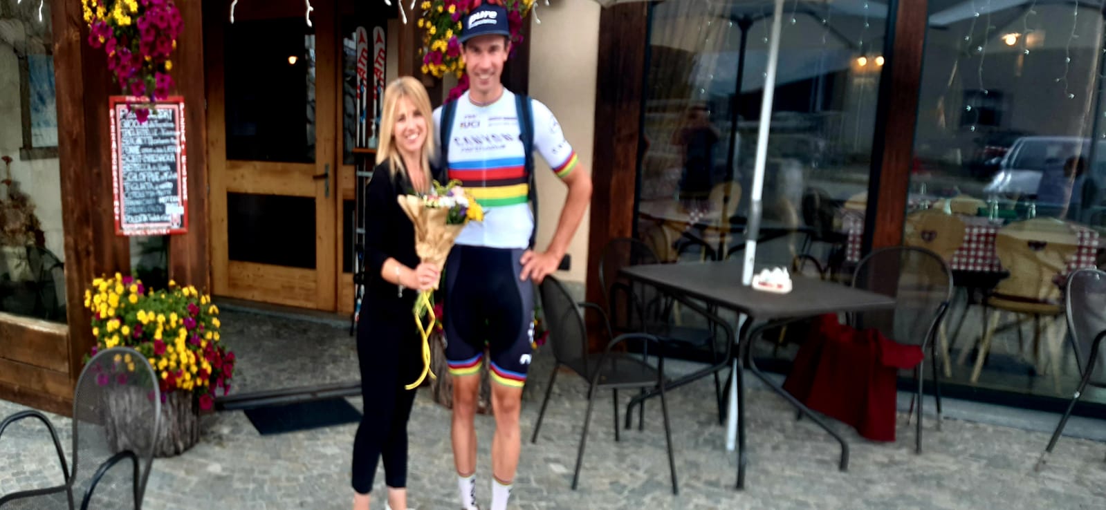 Un ciclista con la maglia arcobaleno è in piedi accanto a una donna che tiene in mano dei fiori fuori da un bar.