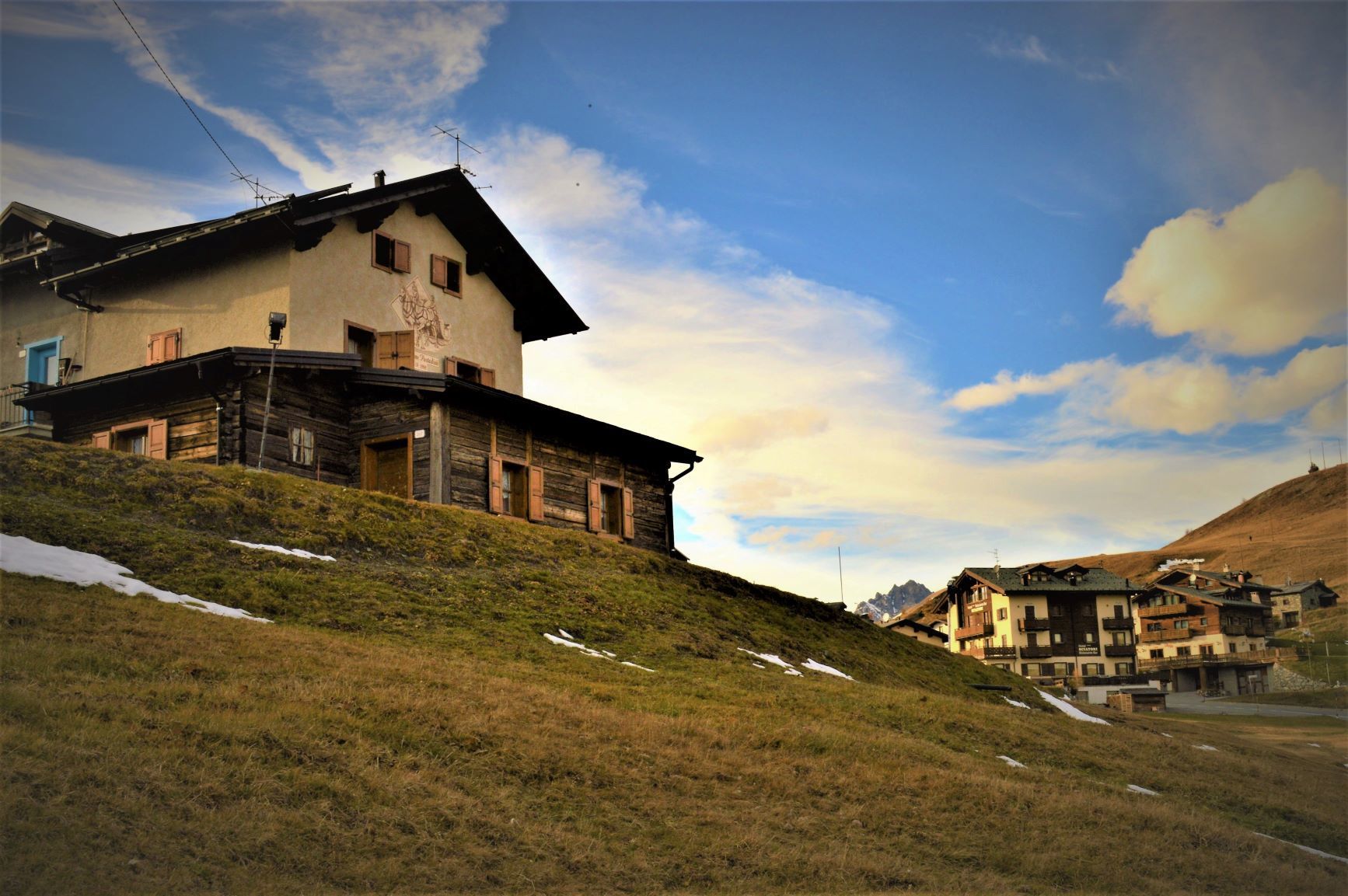 Un edificio a due piani su una collina erbosa; altri edifici in lontananza sotto un cielo azzurro con nuvole.