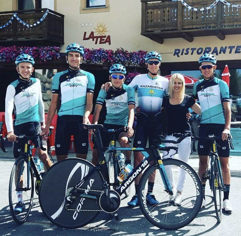 Ciclisti con maglie e caschi color verde acqua posano con una donna davanti a un ristorante.
