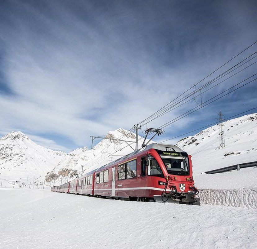 Un treno rosso viaggia attraverso montagne innevate sotto un cielo azzurro.