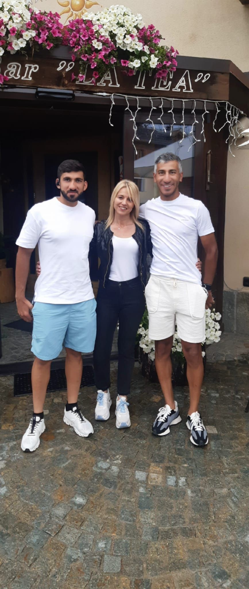 Tre persone posano davanti a un edificio con dei fiori. Due uomini indossano camicie bianche; una donna indossa giacca e pantaloni neri.