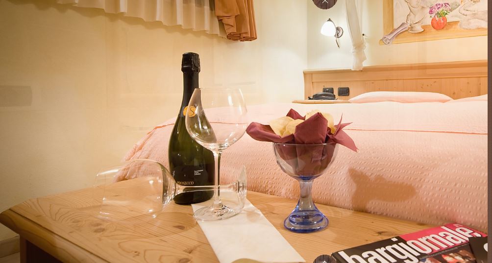 Bottiglia di champagne e bicchieri su un comodino, con un letto e dei fiori sullo sfondo.