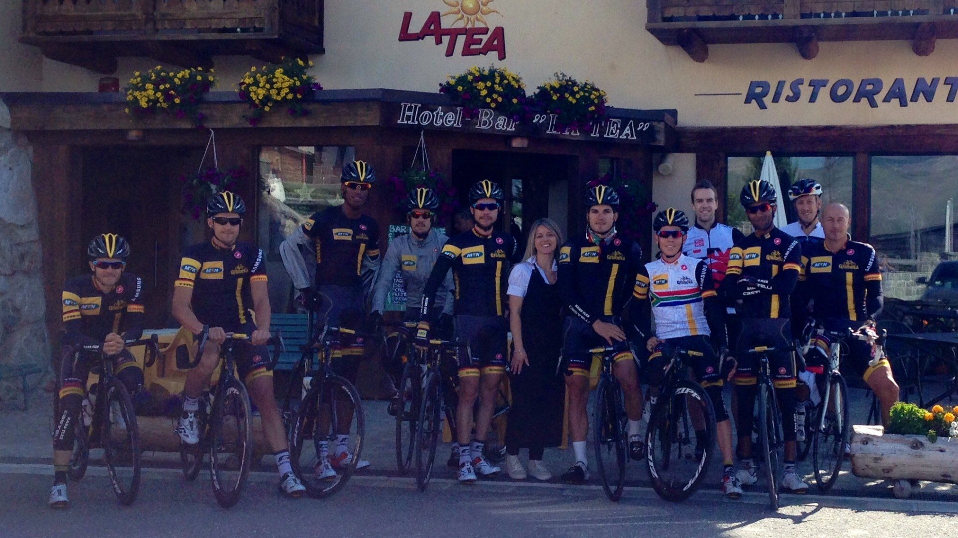Ciclisti in divisa nera e gialla posano davanti a un ristorante, alcuni sorridenti, davanti al cartello
