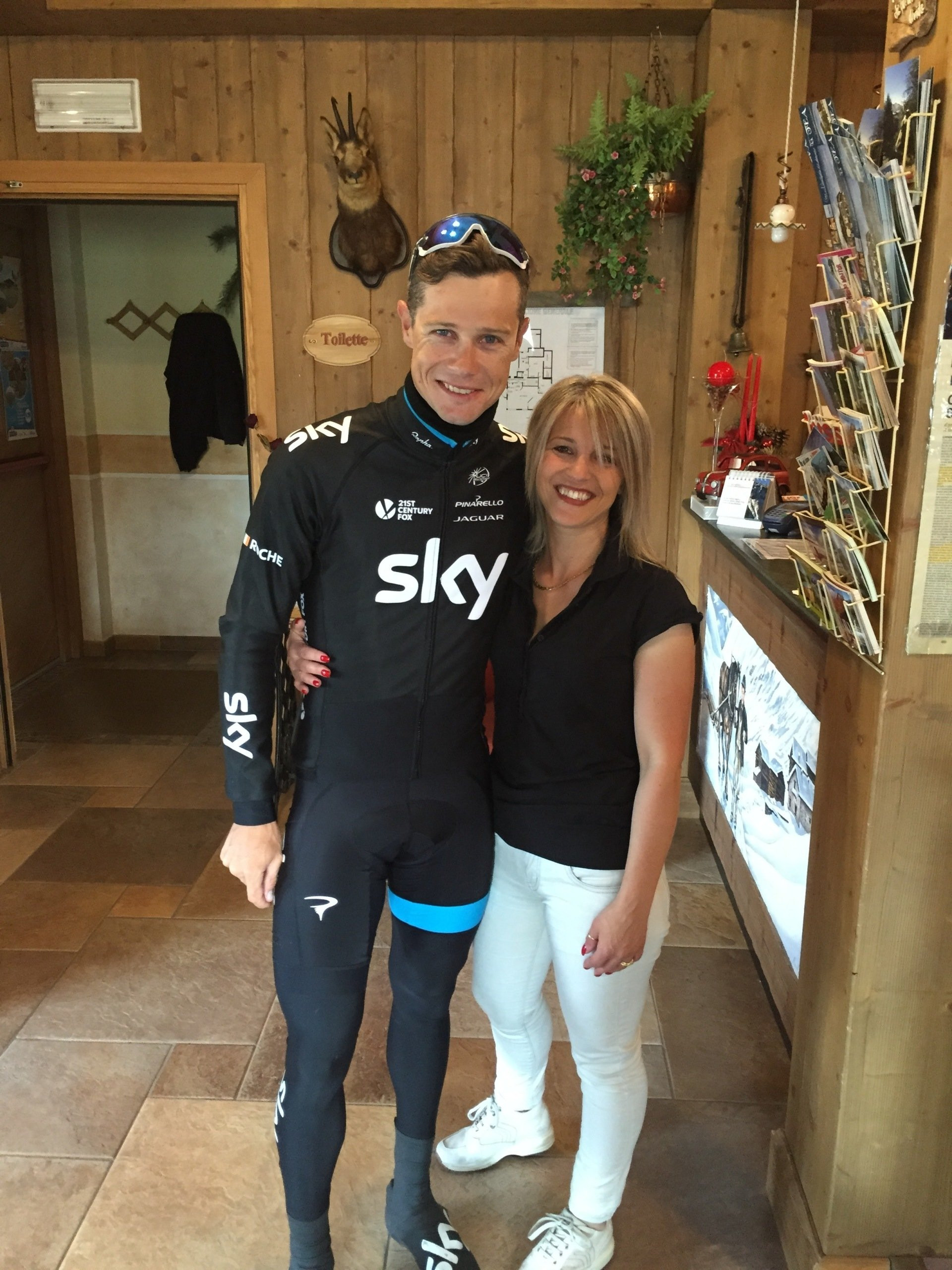 Un ciclista con la maglia nera della Sky posa con una donna sorridente in jeans bianchi davanti a un bancone di legno.