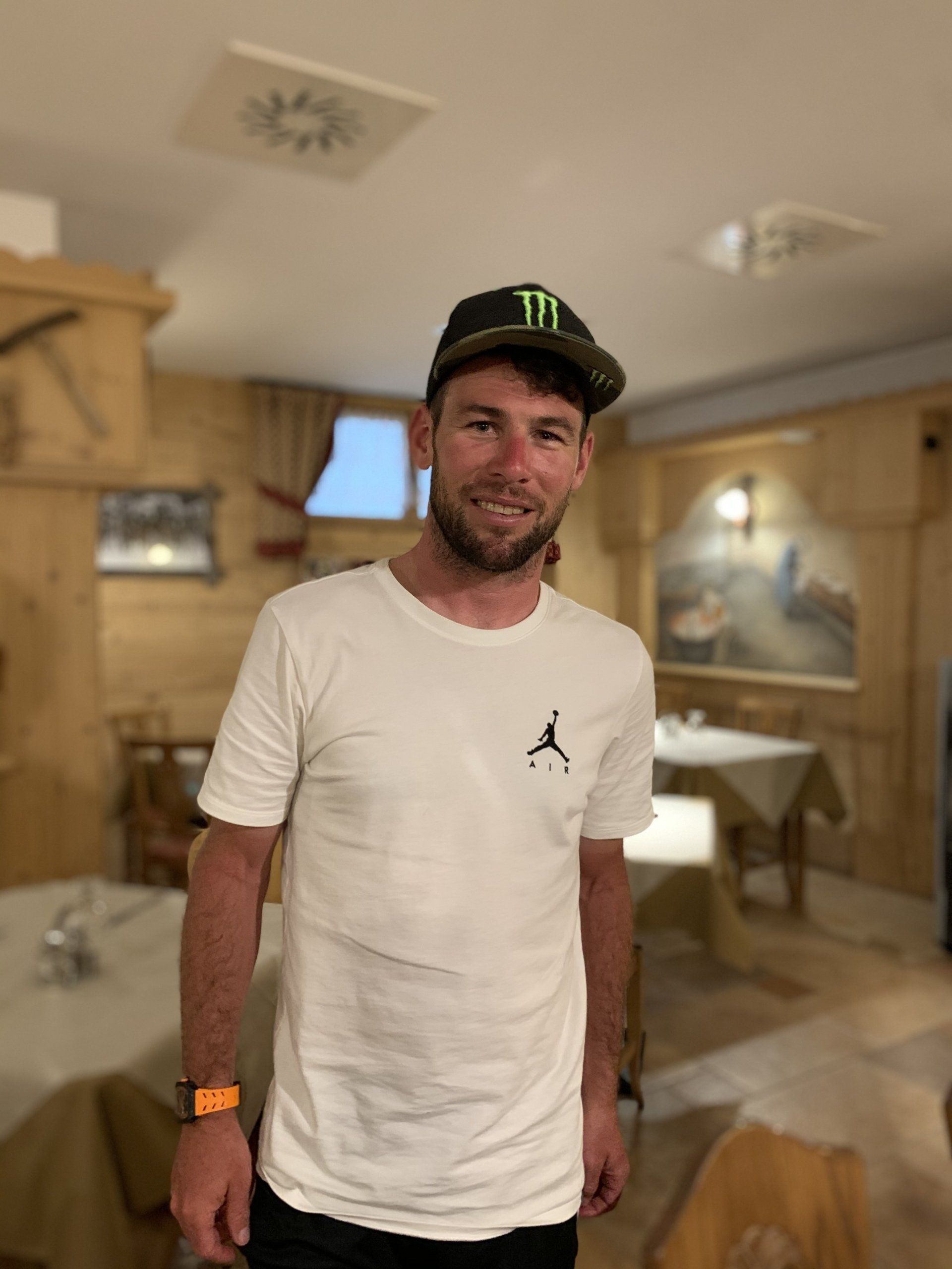 Uomo con maglietta bianca e cappellino Monster Energy, sorridente in un ristorante con pannelli in legno.