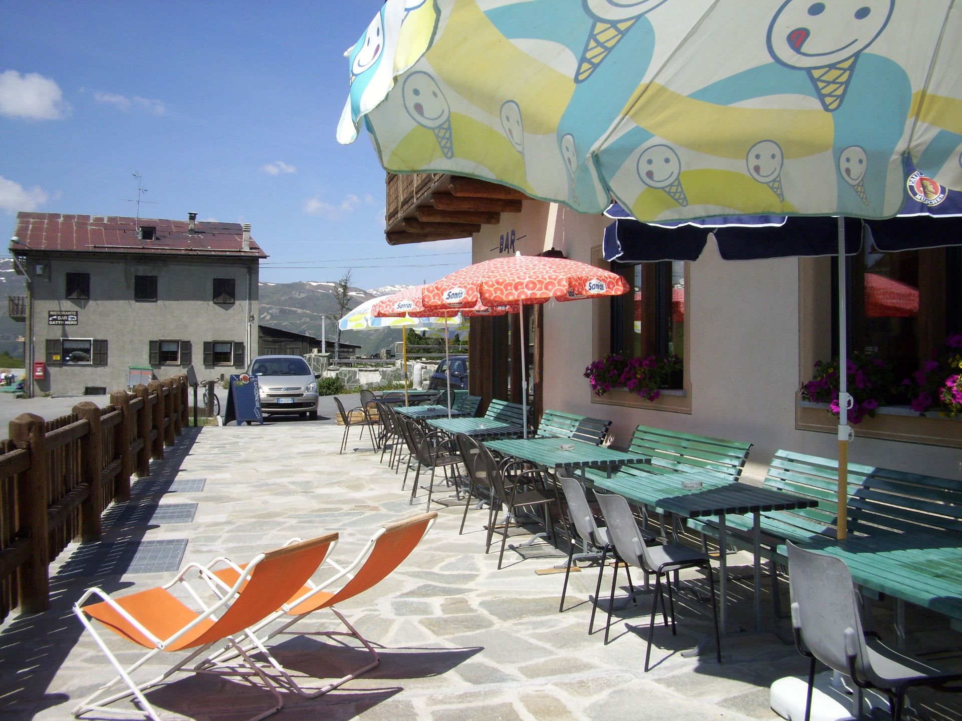 Bar all'aperto con tavoli, ombrelloni e sedie a sdraio su un patio in pietra. Montagne ed edifici sullo sfondo.