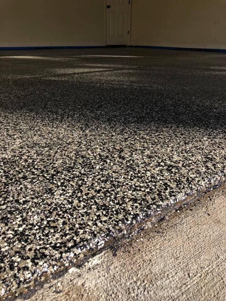 Close-up of a finished garage floor with a speckled, gray, and black epoxy coating. The floor's edge is near a concrete wall.