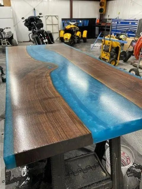 A wooden table with a river of blue resin running through the middle, indoors with motorcycles and equipment in the background.