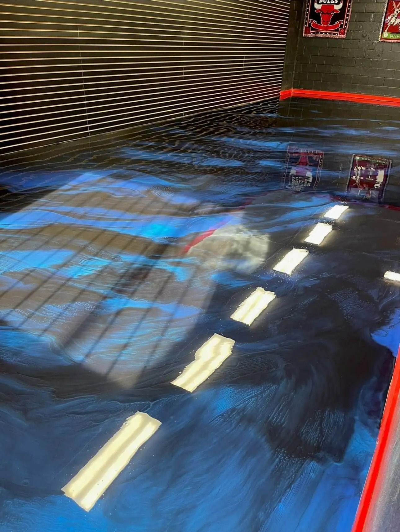 Blue and black epoxy floor with white stripes, reflecting light and the shadow of a grid. Red accents are visible.