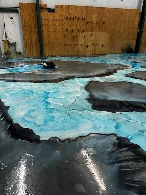 Epoxy floor art with a blue river-like design surrounded by dark gray, on a concrete floor in a workshop.