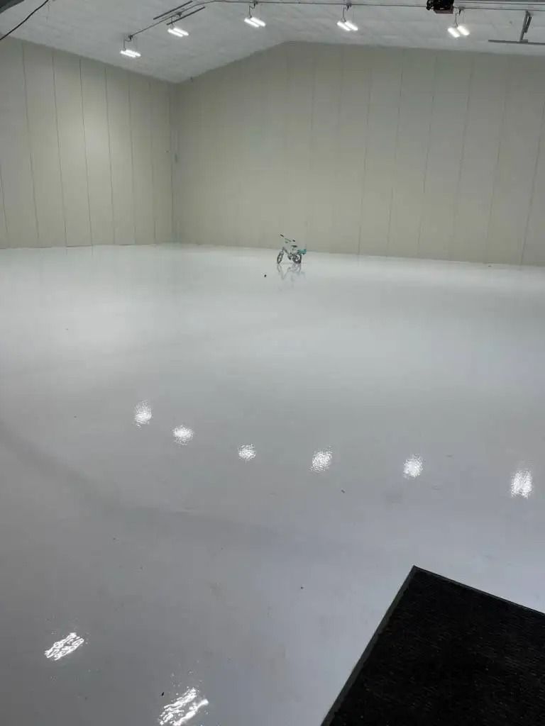 A small, silver robot stands in the center of a vast, empty white room. The room's walls and floor are also white.