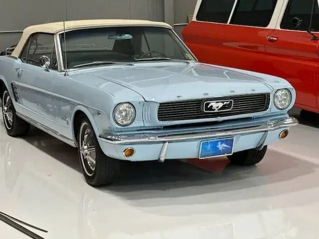 Light blue 1965 Ford Mustang convertible with a white top parked indoors, with a red vehicle partially visible in the background.