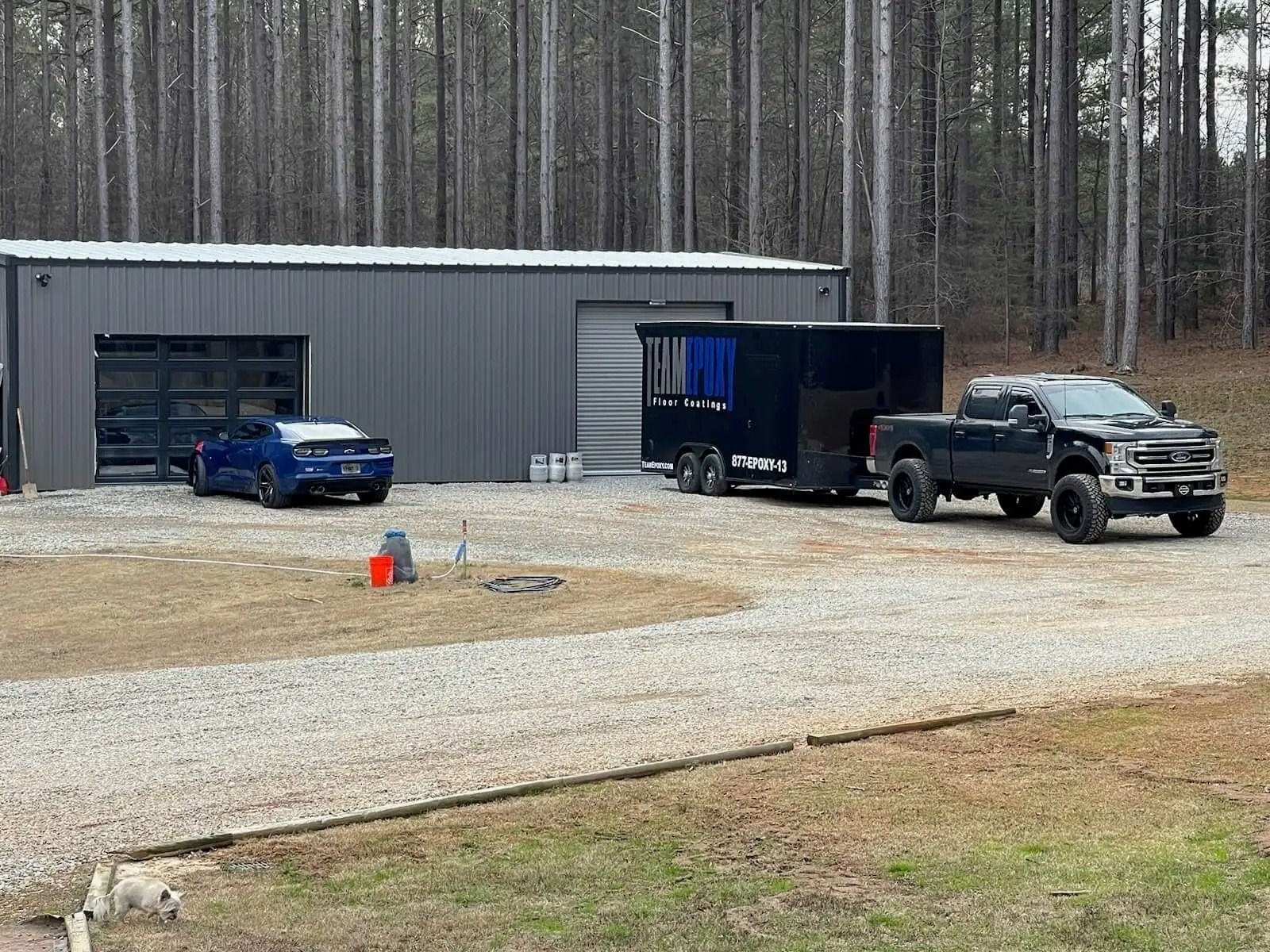 A blue sports car and a black truck with a trailer sit outside a gray metal building in a wooded area.