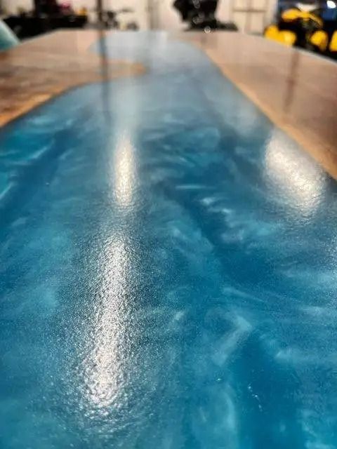 Close-up of a wooden table with a vibrant, ocean-blue resin center. The resin appears swirly and translucent.