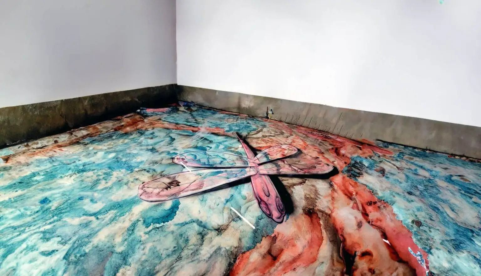 Floor covered in blue and red paint with a butterfly shape in the center, against a white wall in a corner.