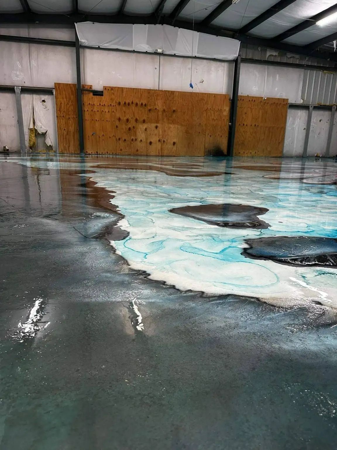 An empty warehouse with a wet, peeling floor that has areas of blue and black color. A wooden barrier is attached to the back wall.