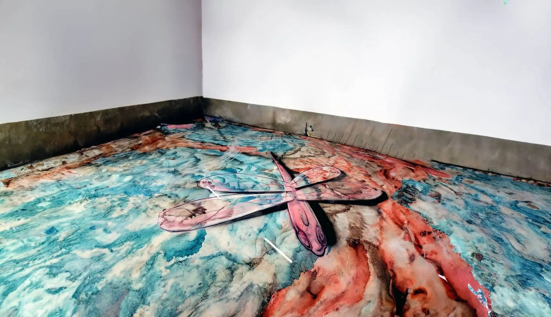 A corner of a room with a floor covered in swirling blue, pink, and red paint. A large dragonfly-like shape lies on the floor.