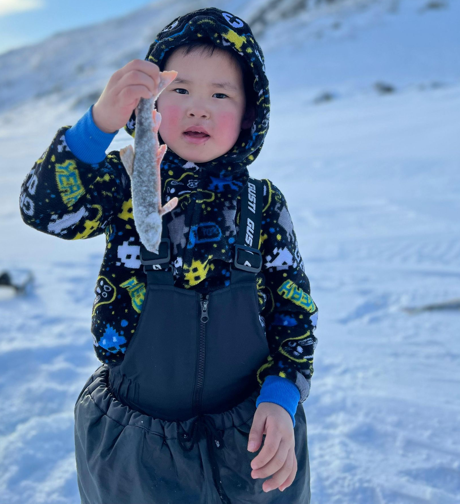 Young child in winter gear holding a fish, snowy landscape in background.